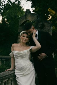 A couple in wedding attire poses romantically by a stone balustrade in a dark, atmospheric garden setting.