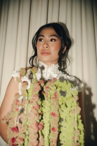 A bride in a high-neck 3D floral lace gown looking away while holding a cascading bouquet of green and pink blooms.