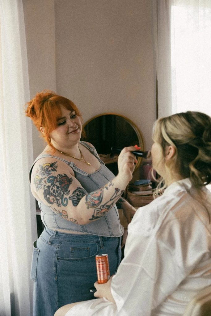 A redhead makeup artist with tattoos applying blush to a blonde bride wearing a white silk robe in a bright room.