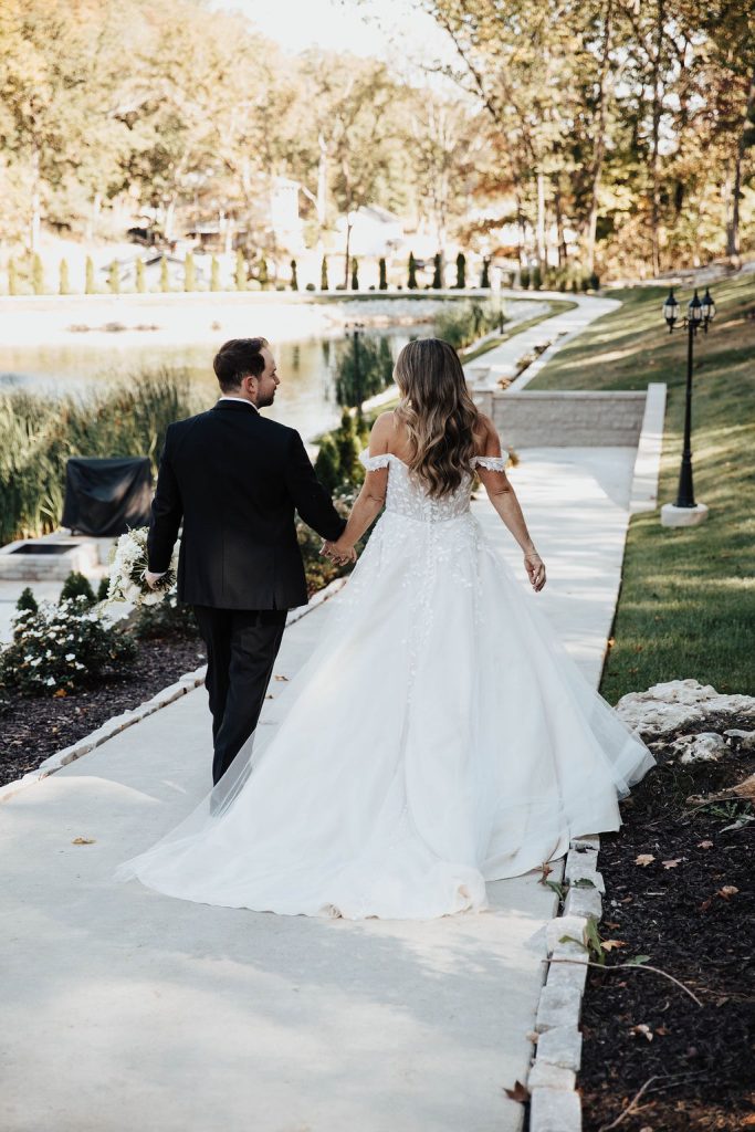 Bride in a Mia Grace gown and groom walking hand-in-hand down a path by the lake at Knotting Hills.