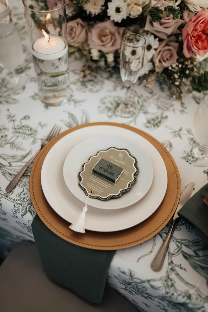 Elegant wedding place setting with a gold charger, sage green napkin, and personalized menu on a floral tablecloth at Knotting Hills.
