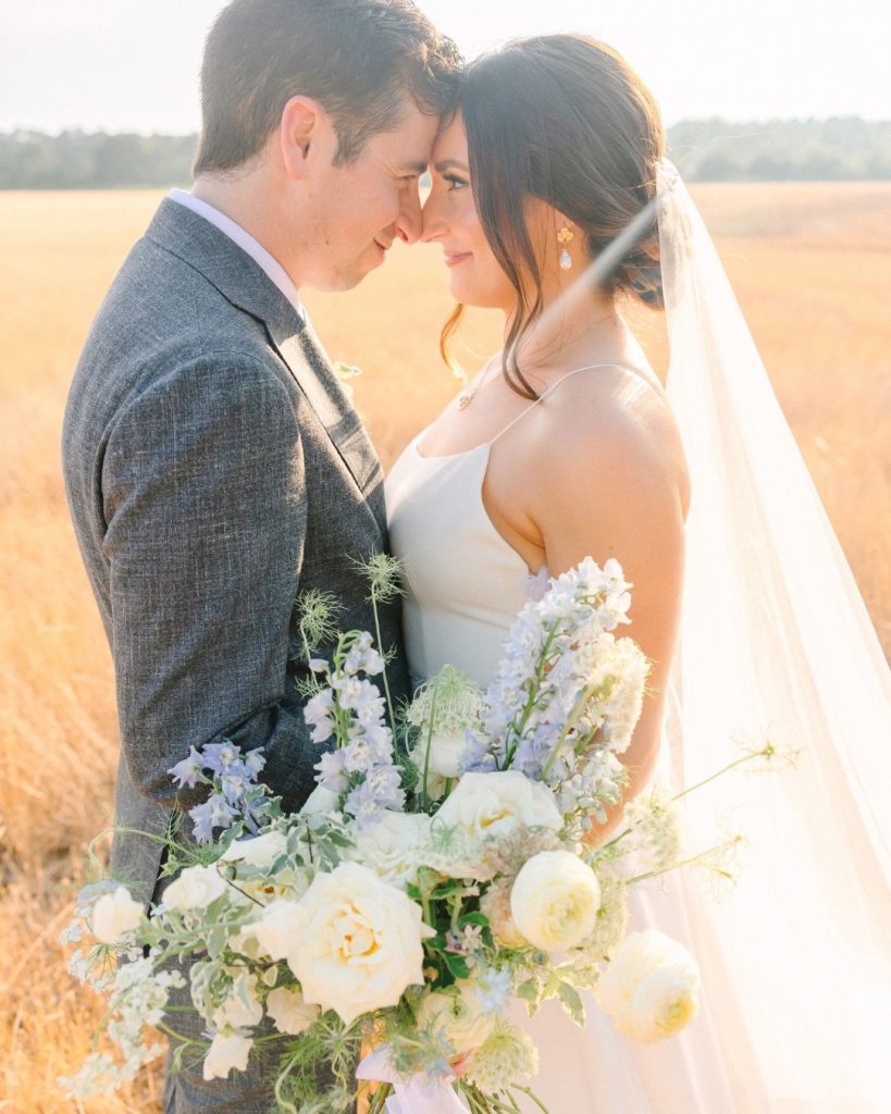 Bride and groom touching foreheads in a sunlit field while the bride holds a loose white-and-pale-blue garden bouquet.