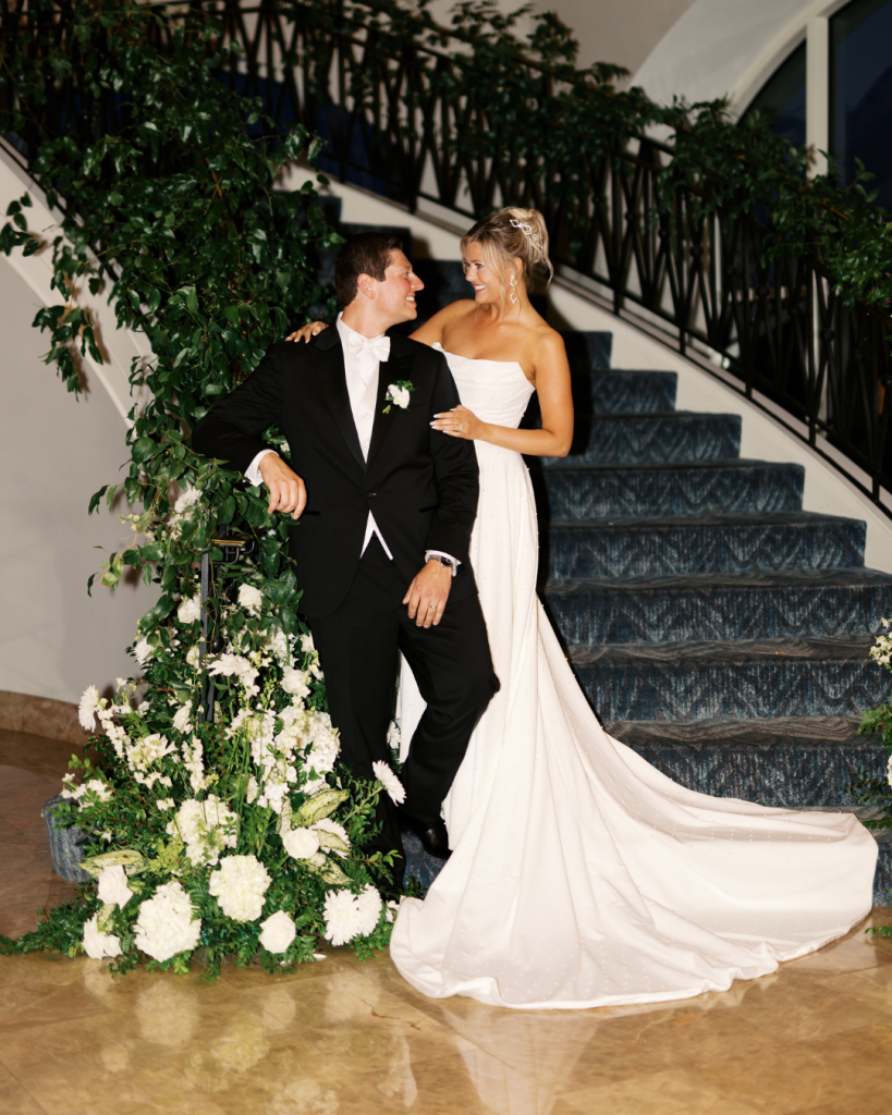 Bride and groom pose on a grand staircase surrounded by white floral installations and greenery at The Ballantyne Hotel.