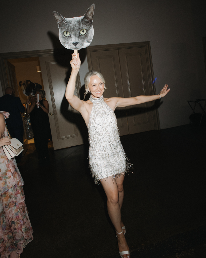Bride dances at her wedding reception wearing a sparkling fringe reception dress while holding a playful cat cutout during a lively celebration at The Ruth.