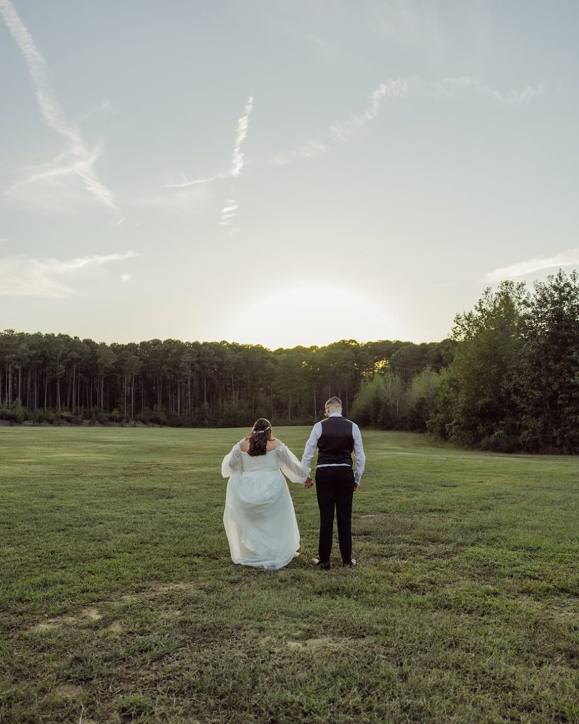 Bride and groom walking hand in hand through an open green field at golden hour, facing a warm sunset sky above a Carolina pine tree line.