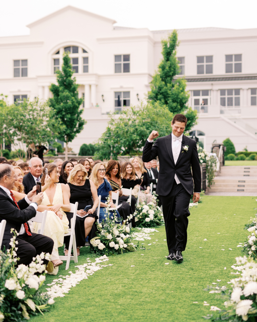 Groom walks down the ceremony aisle smiling as guests look on during an outdoor wedding at The Ballantyne Hotel.
