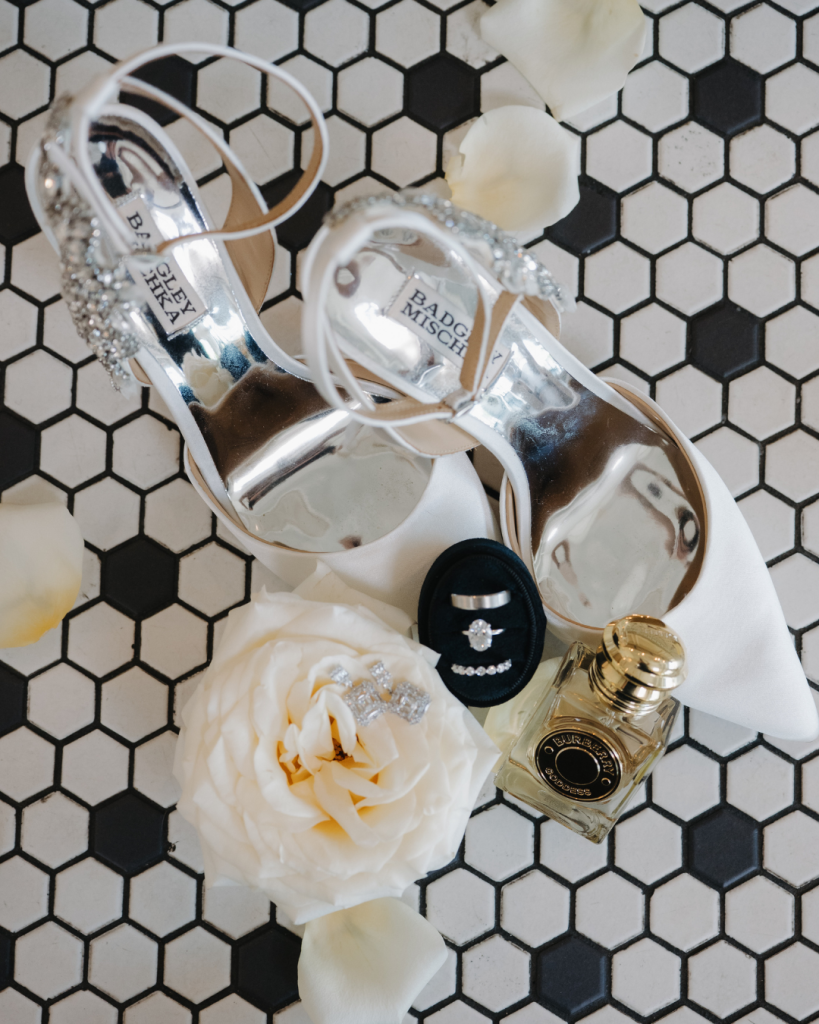 Bridal detail flat lay featuring white Badgley Mischka heels, wedding rings, perfume, earrings, white rose petals, and bridal accessories styled on black and white tile for a Charlotte wedding.