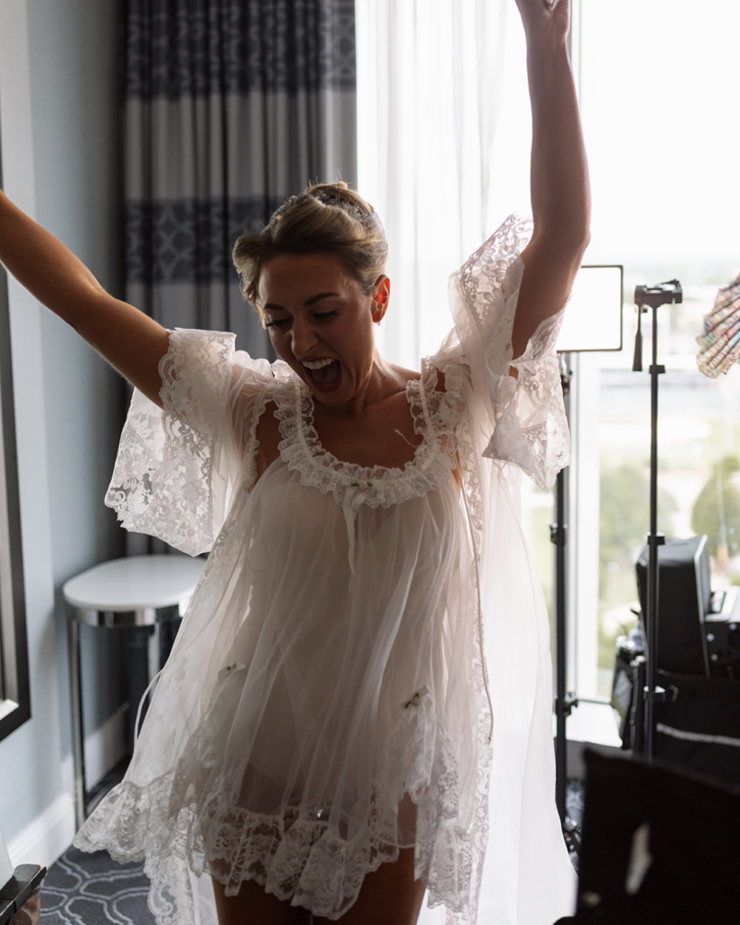 Bride Valerie celebrates while getting ready in a lace trimmed bridal robe inside a Charlotte hotel suite before her wedding day.