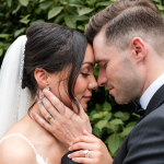 Close-up of bride and groom embracing forehead-to-forehead, her hand on his face showing her engagement ring and wedding band, surrounded by greenery.