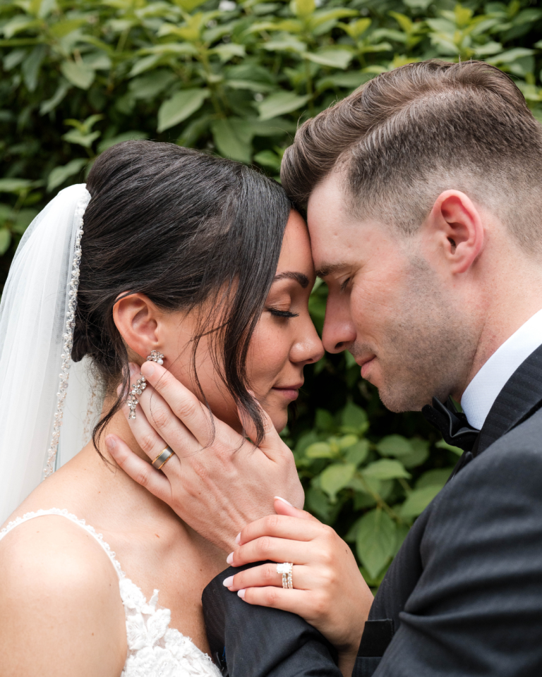 Close-up of bride and groom embracing forehead-to-forehead, her hand on his face showing her engagement ring and wedding band, surrounded by greenery.