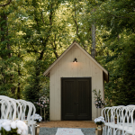 White ceremony chapel with dark double doors flanked by pink and white floral arrangements, viewed down a white aisle runner lined with empty chairs beneath a leafy tree canopy.