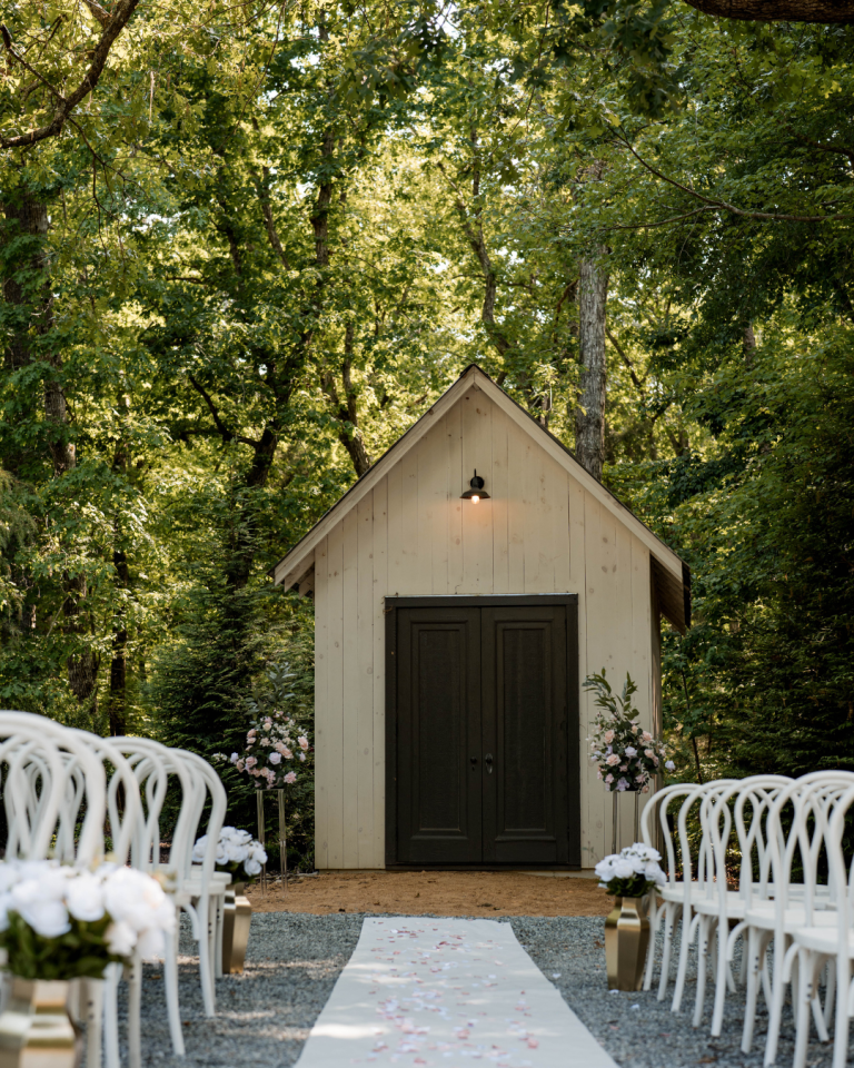 White ceremony chapel with dark double doors flanked by pink and white floral arrangements, viewed down a white aisle runner lined with empty chairs beneath a leafy tree canopy.