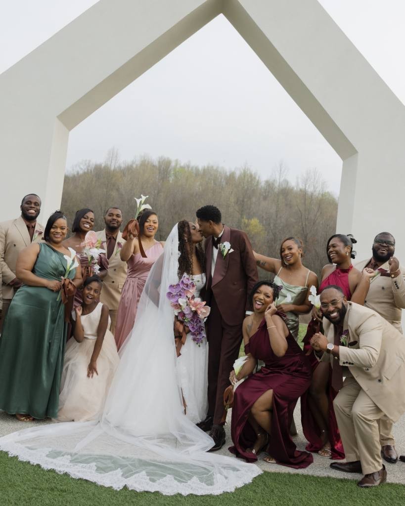Bride and groom kiss beneath the signature white ceremony structure at The Lumen House surrounded by their colorful wedding party.