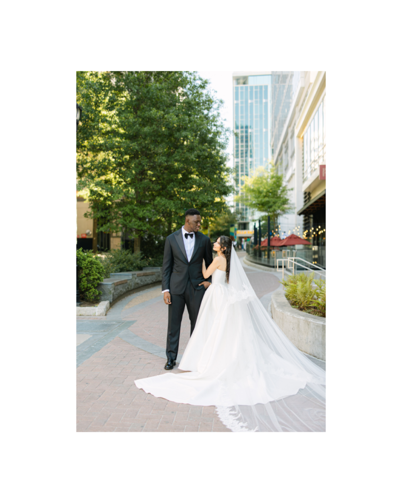 Bride and groom walk together through Uptown Charlotte with the bride's cathedral veil trailing behind during editorial wedding portraits.