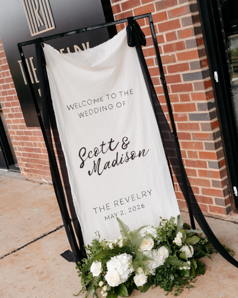 Modern black and white wedding welcome sign for Madison and Scott displayed outside The Revelry with white floral arrangements and black draping.