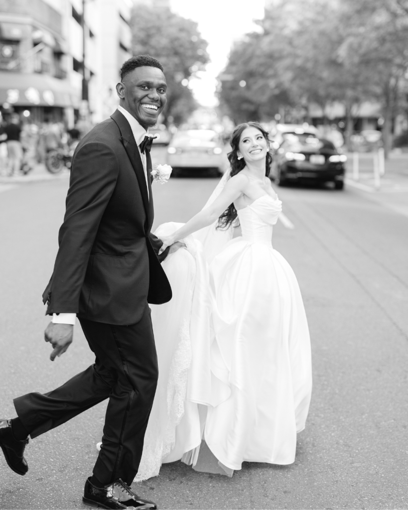 Black and white wedding portrait of the bride and groom smiling while crossing a Charlotte street together after their ceremony.