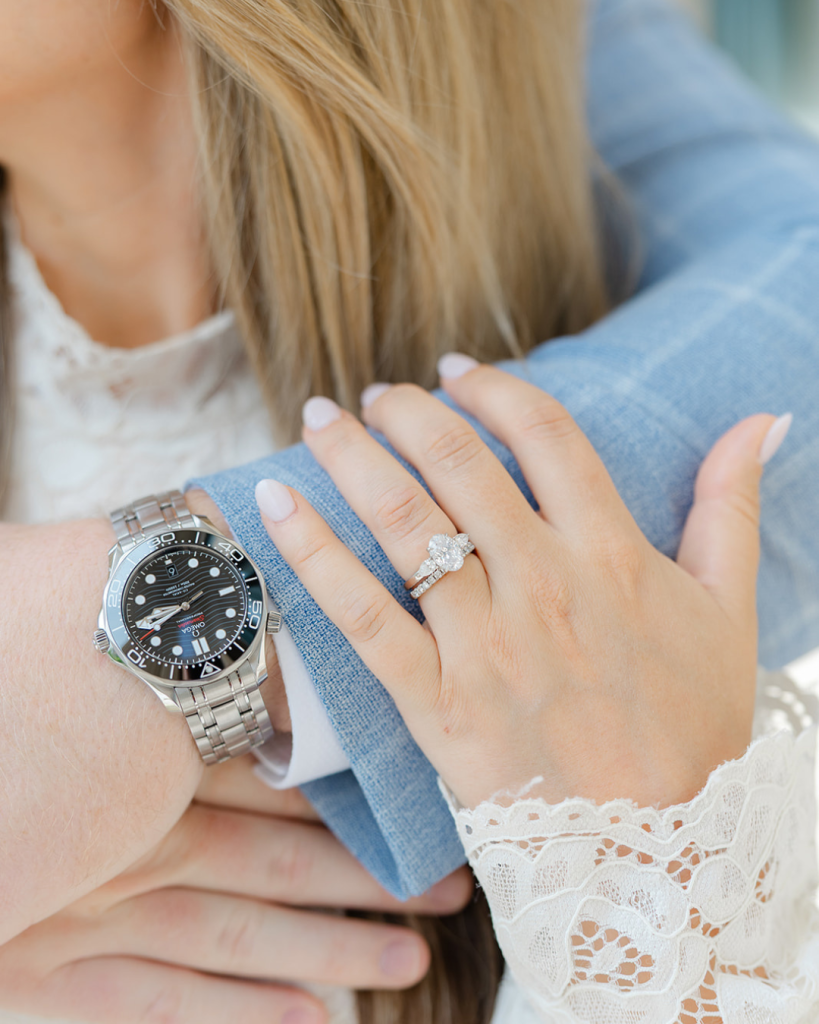 Close up of Audrey's diamond engagement ring and wedding band as she rests her hand on Steve's light blue suit jacket. A luxury watch is visible on his wrist.