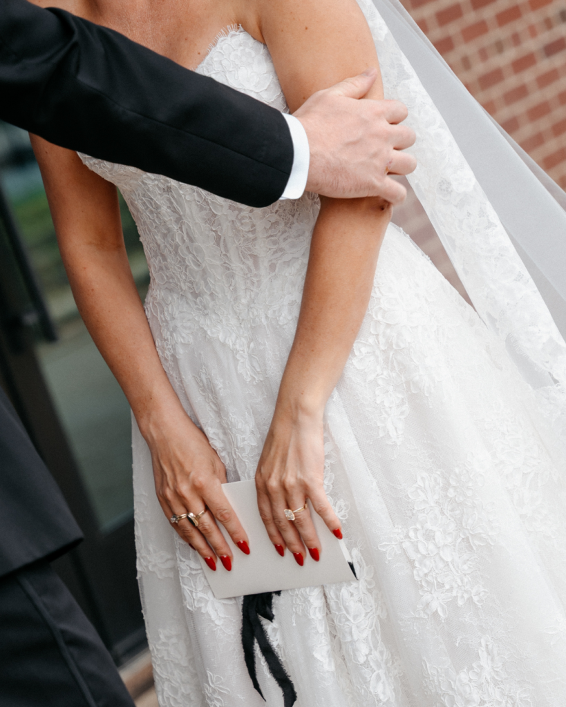 Detail photo of bride's lace wedding gown, engagement ring, red manicure, and black ribbon accents during wedding portraits.