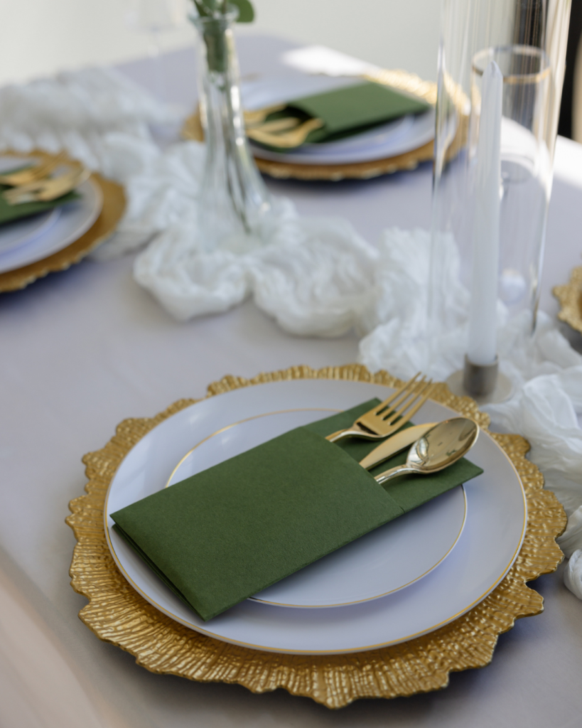 Elegant wedding reception place setting with gold charger plate, white china, green napkin, and modern tablescape details.