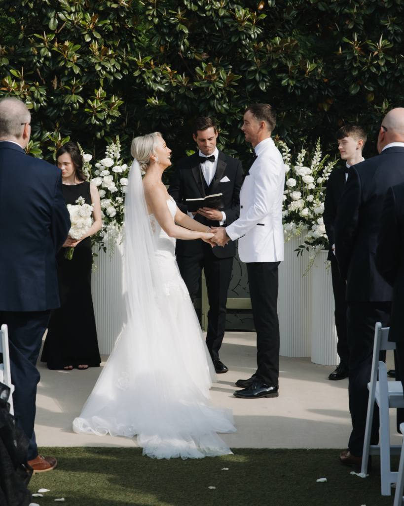Danielle and Ryan exchange vows during their outdoor ceremony surrounded by white floral arrangements and loved ones at The Ruth in Charlotte, North Carolina.