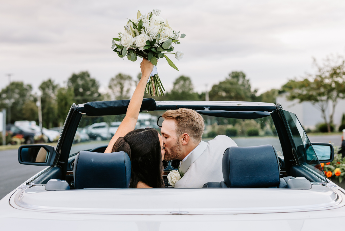 Bride and groom kissing in a vintage convertible as the bride raises her bouquet during their wedding exit.