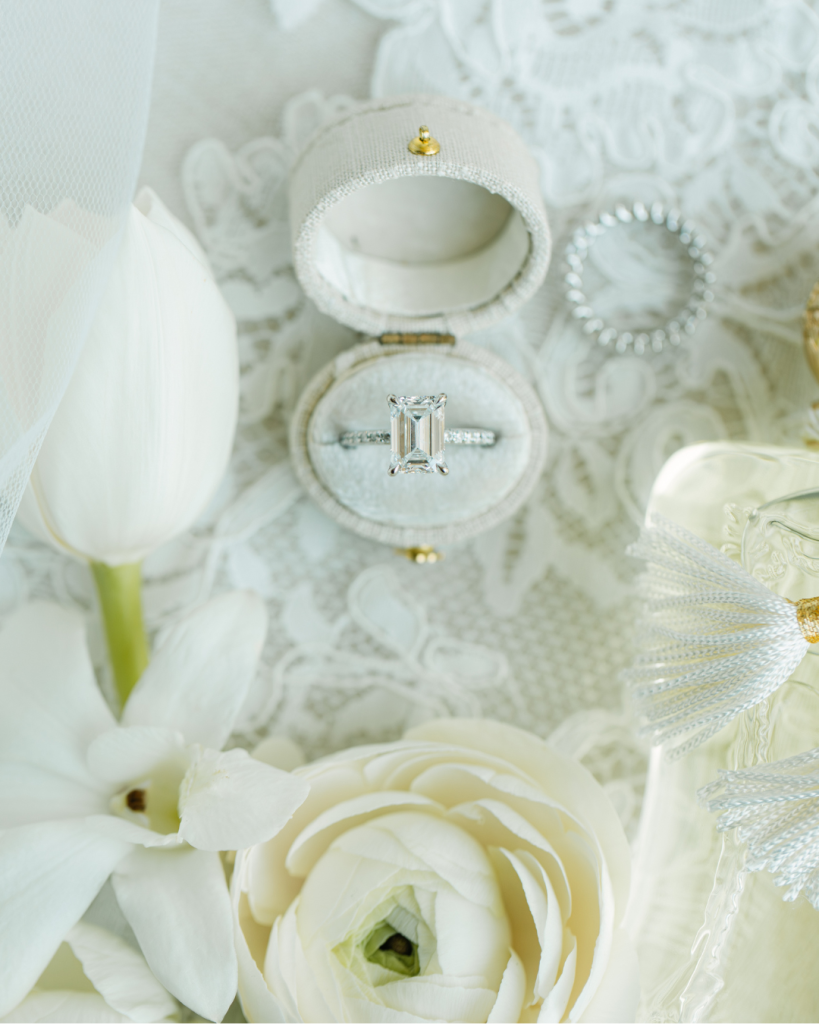 Close up of an emerald cut engagement ring styled with white flowers, lace, perfume, and bridal accessories for wedding detail photography in Charlotte.