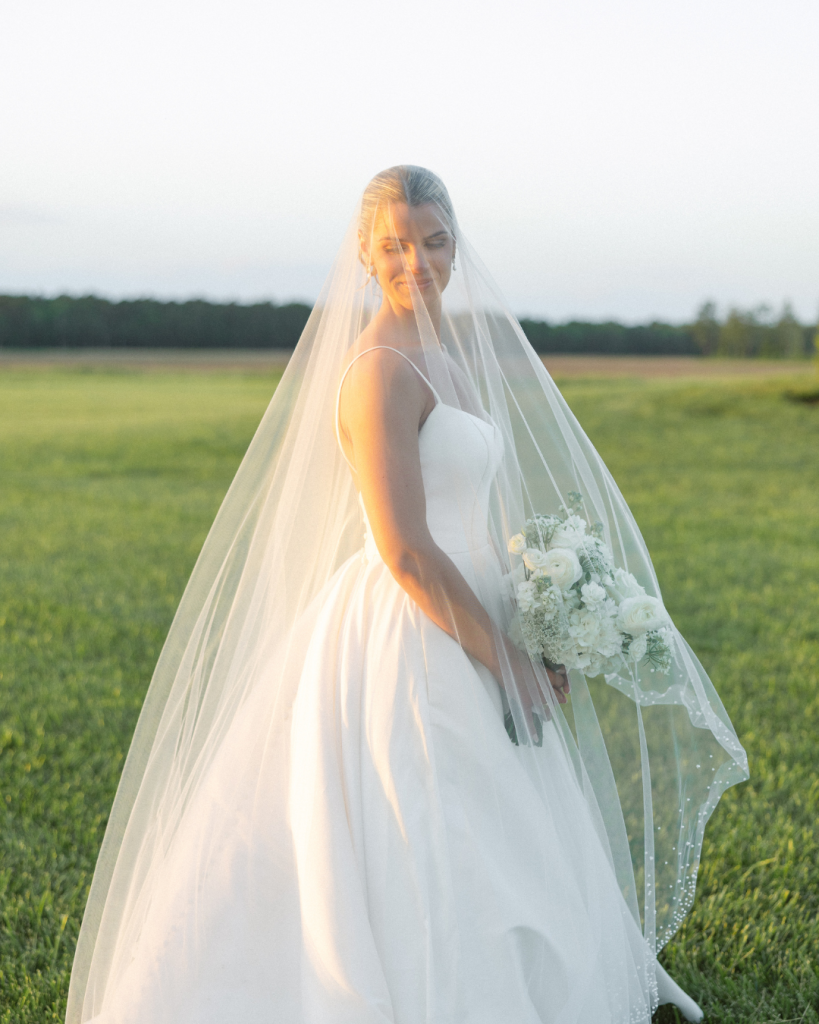 Bride standing beneath her veil in golden evening light holding a white floral bouquet during a Maryland Eastern Shore wedding.