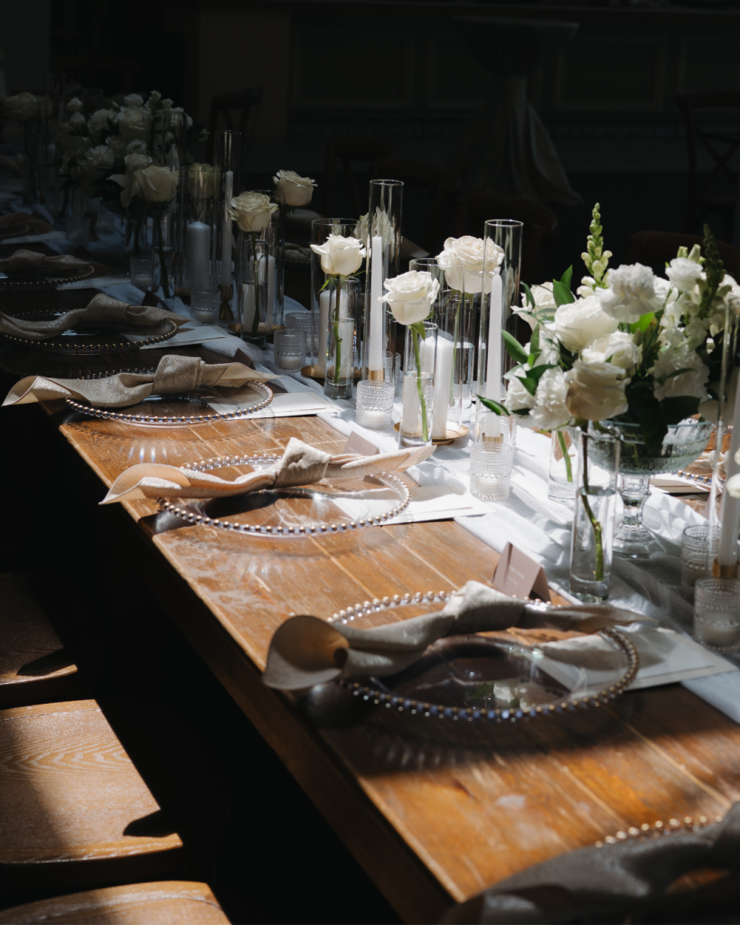 Elegant reception tablescape featuring candlelight, white roses, glass cylinders, and refined place settings styled by SIPS Rentals for a romantic Charlotte wedding.