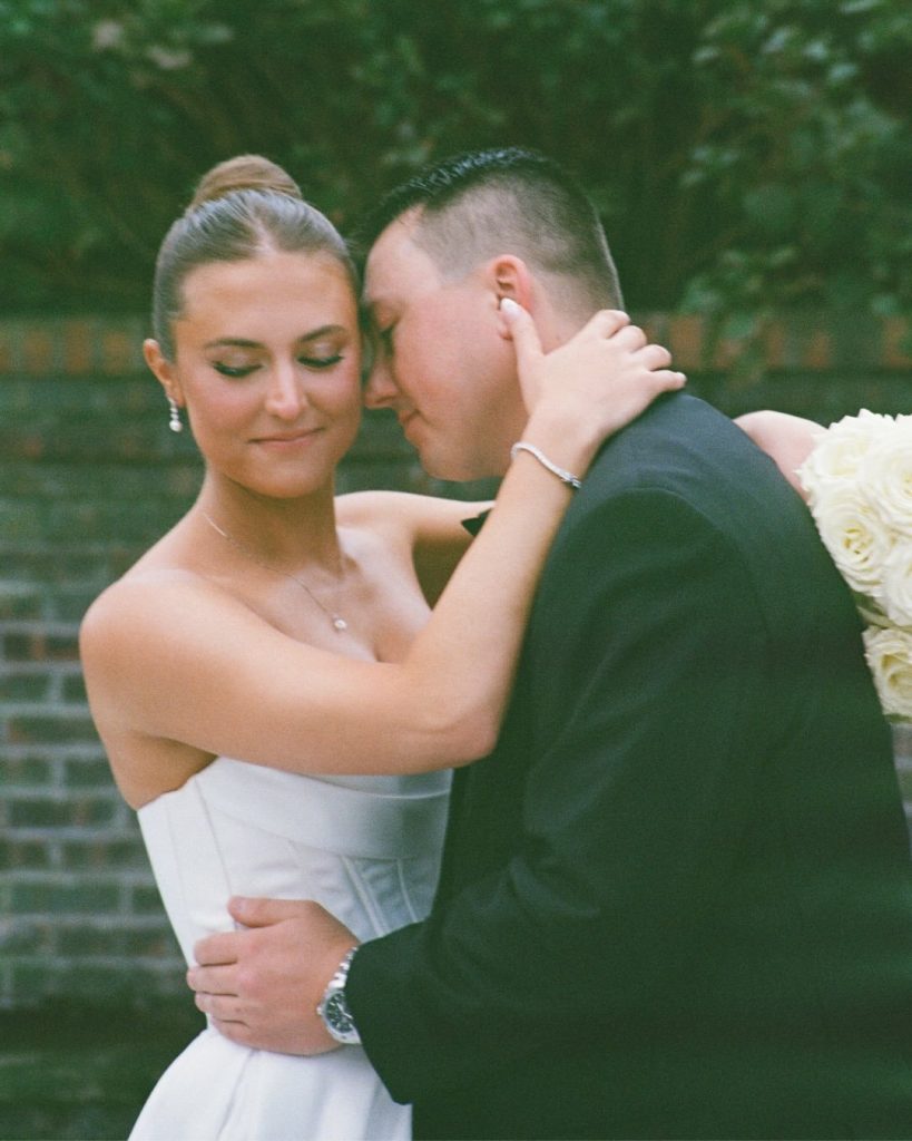 : Bride and groom in an intimate embrace outdoors, foreheads touching, with greenery and a brick wall in the background.