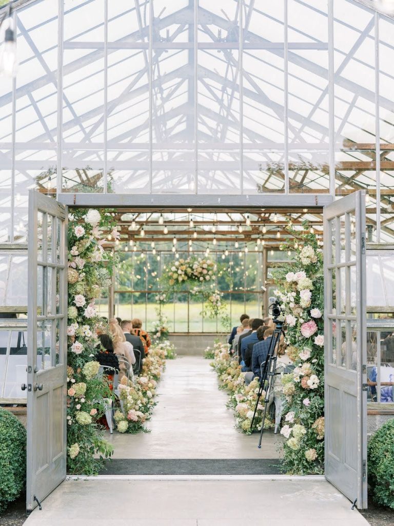 Guests seated for a wedding ceremony inside a glass greenhouse venue with lush floral installations lining the aisle and ceremony arch.