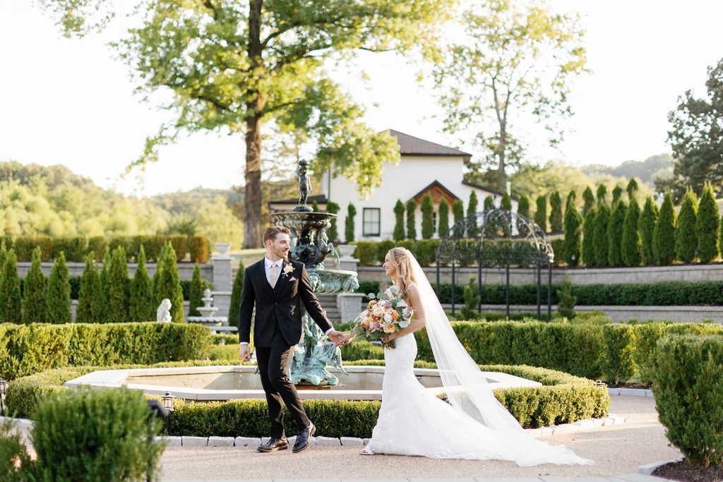 Bride and groom holding hands in front of an ornate fountain at Knotting Hills Wedding Venue + Resort.