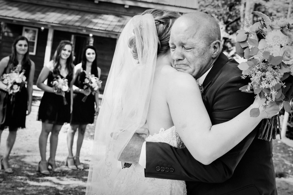 Bride sharing an emotional first look hug with her father before the wedding ceremony while bridesmaids watch in the background.