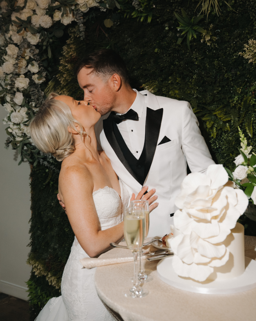 Bride and groom share a romantic kiss beside their elegant white wedding cake surrounded by lush greenery and white florals during their reception at The Ruth in Charlotte, North Carolina.