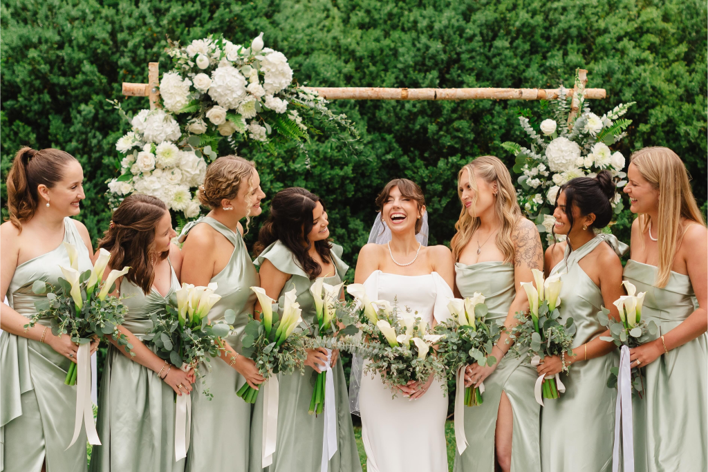 bride laughing with bridesmaids at outdoor wedding altar wearing sage dresses and calla lily bouquet