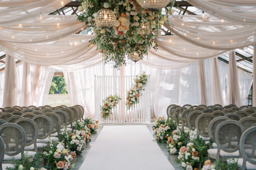 Elegant wedding ceremony setup inside a glass conservatory featuring soft white draping across the ceiling, lush floral arrangements in blush, peach, ivory, and green tones lining the aisle, and a romantic floral installation above the ceremony altar.