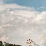 Groom lifting his bride on a grassy hill beneath a dramatic blue and cloudy sky.