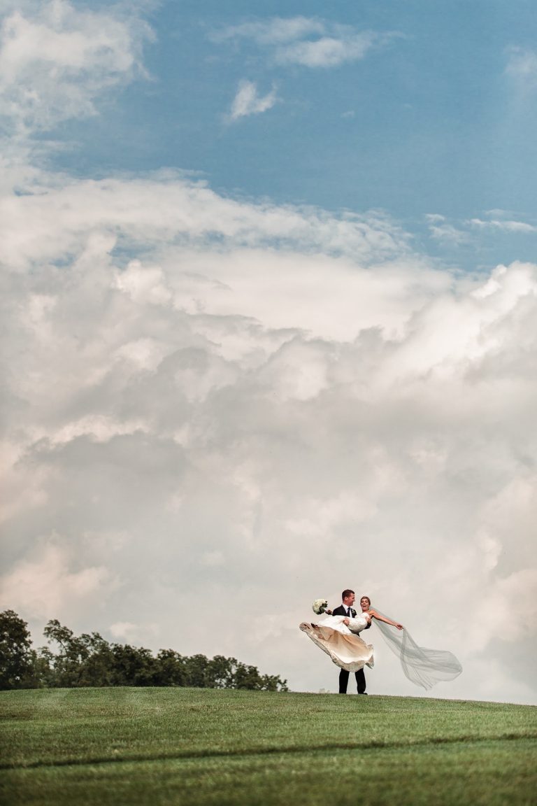 Groom lifting his bride on a grassy hill beneath a dramatic blue and cloudy sky.