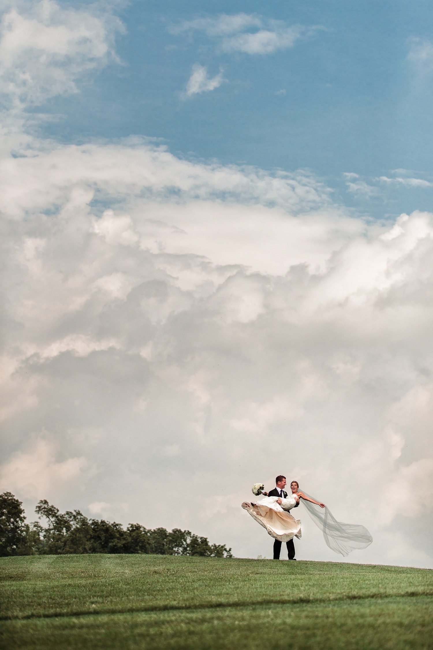 Groom lifting his bride on a grassy hill beneath a dramatic blue and cloudy sky.