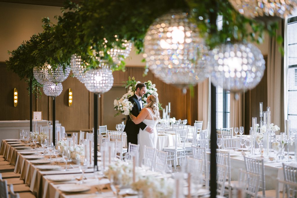 Bride and groom sharing a romantic moment in a modern wedding reception space surrounded by white floral arrangements, crystal chandeliers, and elegant tablescapes with greenery overhead at a Columbus, Ohio wedding.