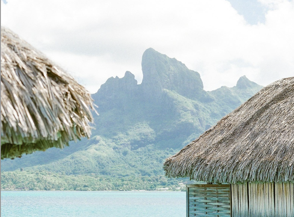 Overwater bungalows with thatched roofs sit along a turquoise lagoon, with lush green mountains rising in the background at a tropical island destination.