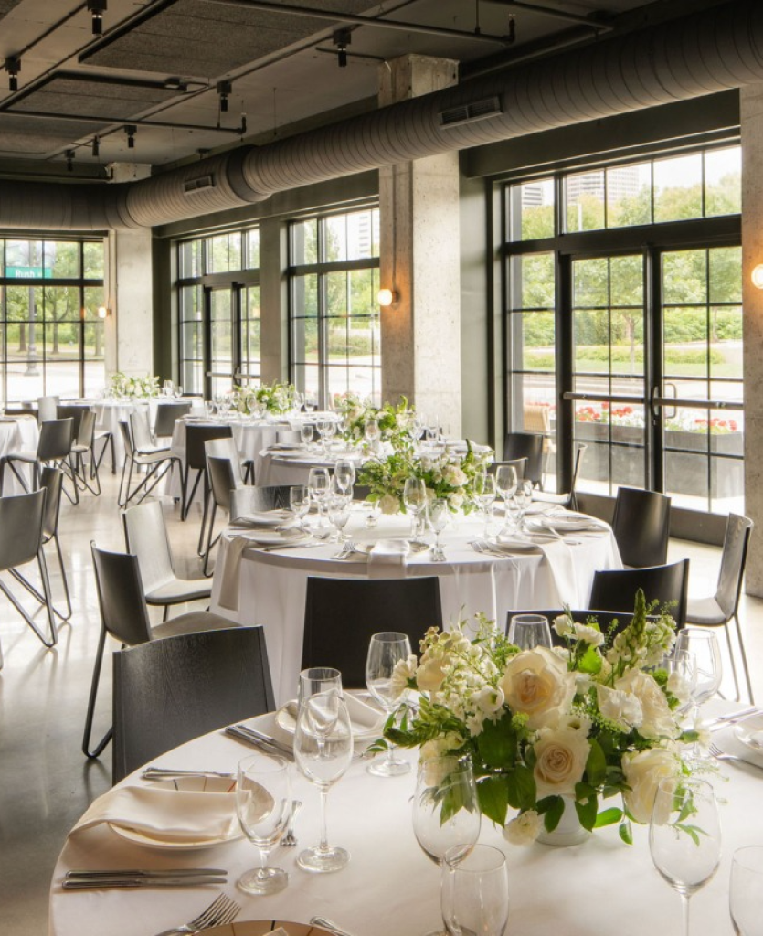 Wedding reception setup in The Belle St. Gallery at The Junto Hotel in Columbus, Ohio, featuring floor-to-ceiling windows, modern industrial design, white floral centerpieces, and elegant round dining tables overlooking downtown Columbus.