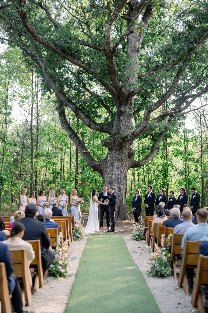 Outdoor wedding ceremony beneath a large mature tree, with the bride and groom exchanging vows surrounded by their wedding party and seated guests. A green aisle lined with colorful floral arrangements leads to the ceremony beneath the tree.
