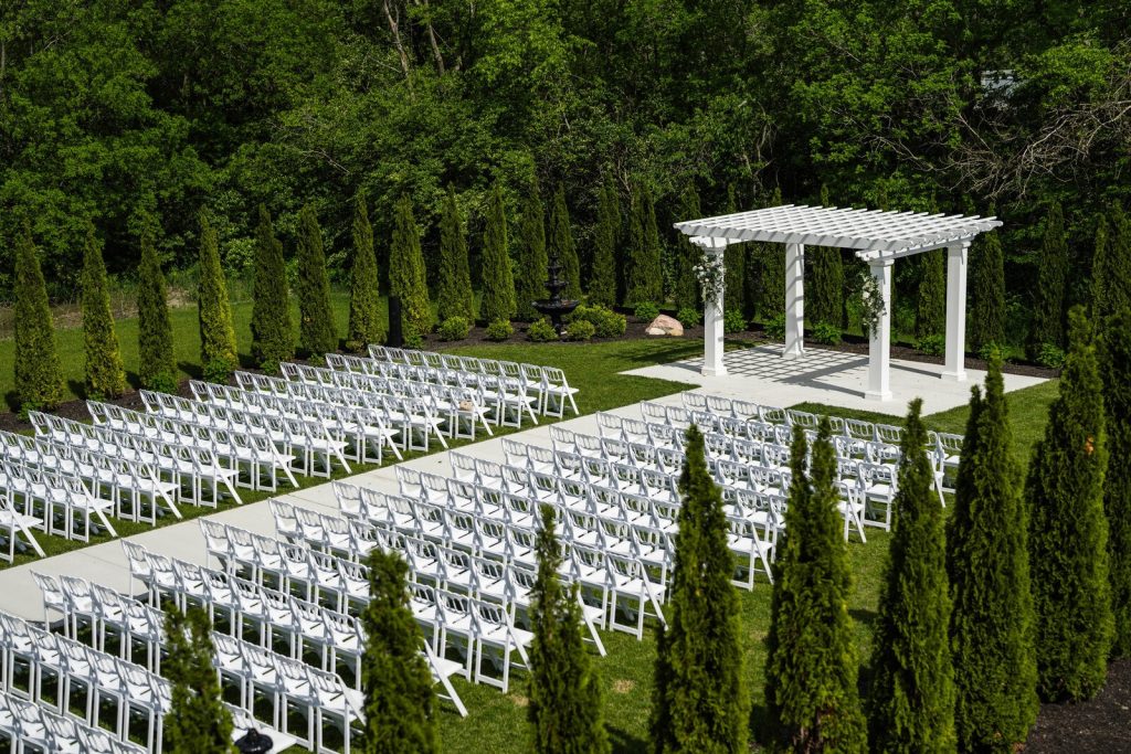 Outdoor wedding ceremony space at White Willow Meadows near Columbus, Ohio, featuring a white pergola altar, rows of white ceremony chairs, manicured landscaping, and tall evergreen trees creating a private garden setting.