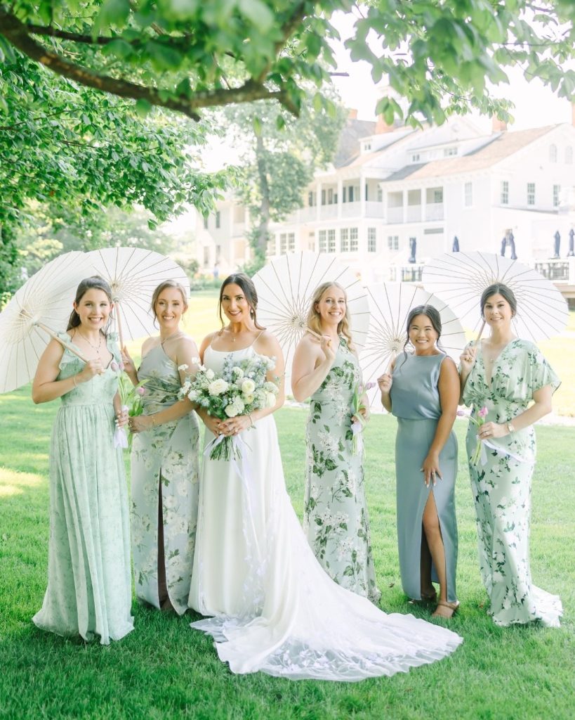 Bride and bridesmaids carrying white parasols in shades of sage and floral-print dresses outside Kent Island Resort.