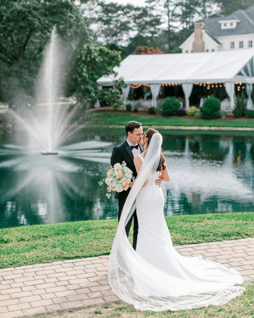 A bride and groom sharing a kiss on a stone brick pathway alongside a pond at an outdoor wedding venue in North Carolina. A large water fountain sprays in the background in front of a white reception tent decorated with bistro lights. The bride wears a fitted lace gown with a long tulle veil flowing behind her, and the groom is in a classic black tuxedo.