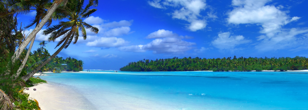 Tropical beach with white sand, crystal-clear turquoise water, and leaning palm trees along the shoreline beneath a bright blue sky with scattered clouds.