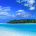 Tropical beach with white sand, crystal-clear turquoise water, and leaning palm trees along the shoreline beneath a bright blue sky with scattered clouds.