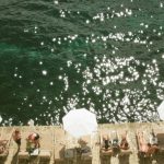 Aerial view of lounge chairs and umbrellas lining a stone pool deck beside sparkling turquoise water, with guests relaxing in the sun.