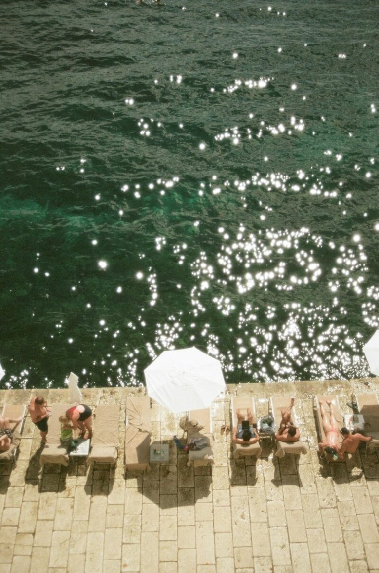 Aerial view of lounge chairs and umbrellas lining a stone pool deck beside sparkling turquoise water, with guests relaxing in the sun.