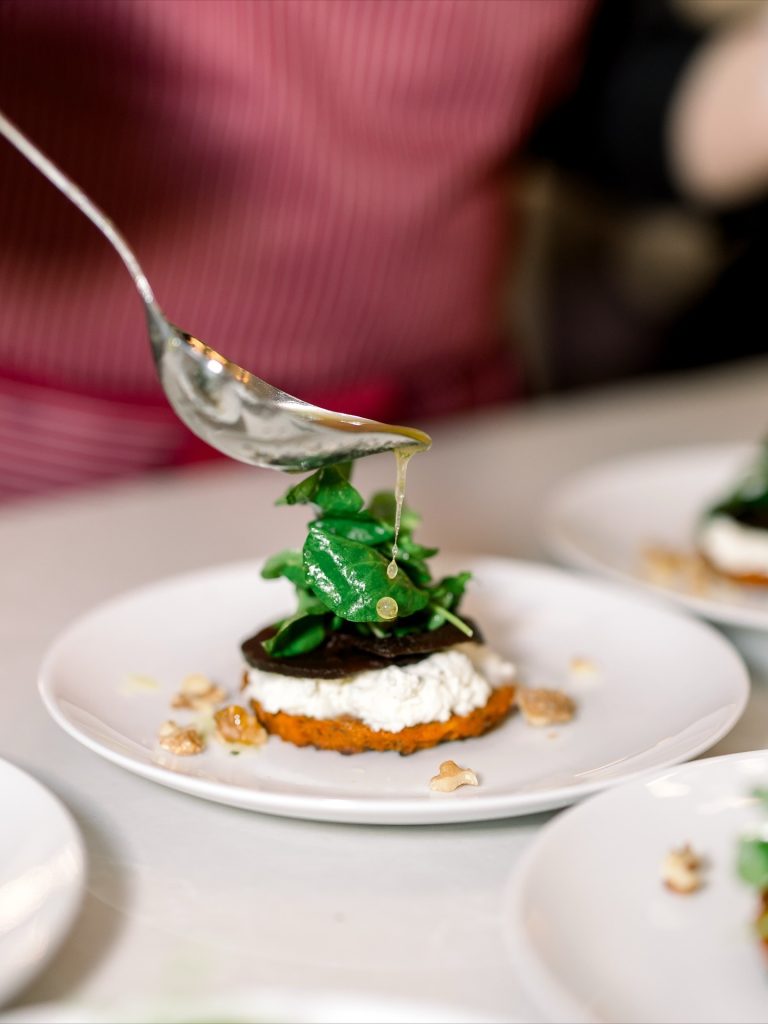 Close-up of a plated appetizer topped with fresh greens as dressing is drizzled from a spoon, featuring layered cheese and vegetables with walnut garnish.